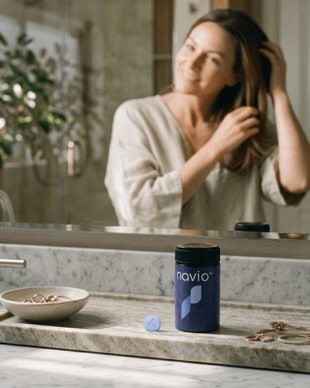 Woman smiling at her reflection in a bathroom mirror, touching her hair, with a Navio bottle on the counter
